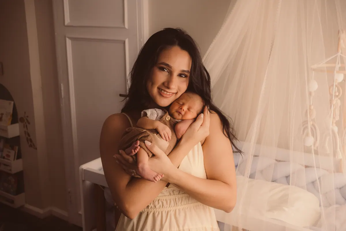 Mãe sorrindo com o bebê recém-nascido no colo, ao lado do berço com dossel